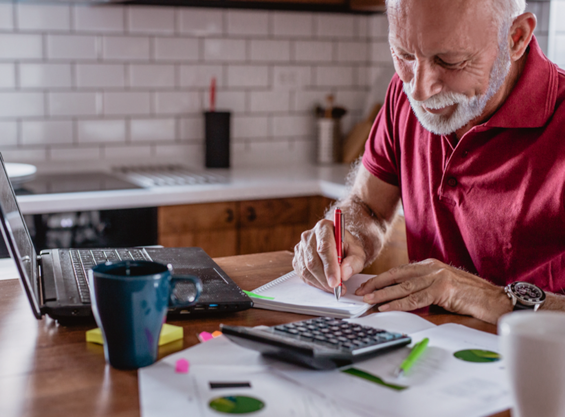 Man writing notes in book with his laptop in front of him